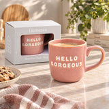 A pink ceramic mug with 'HELLO GORGEOUS' printed in white sits on a marble countertop next to its white packaging box. The scene is styled with a striped cloth, cookies, and a potted plant in the background.